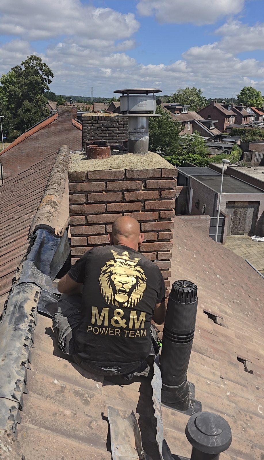 Teamlid met het M&M-logo op zijn shirt werkt bij een bakstenen schoorsteen op een pannendak