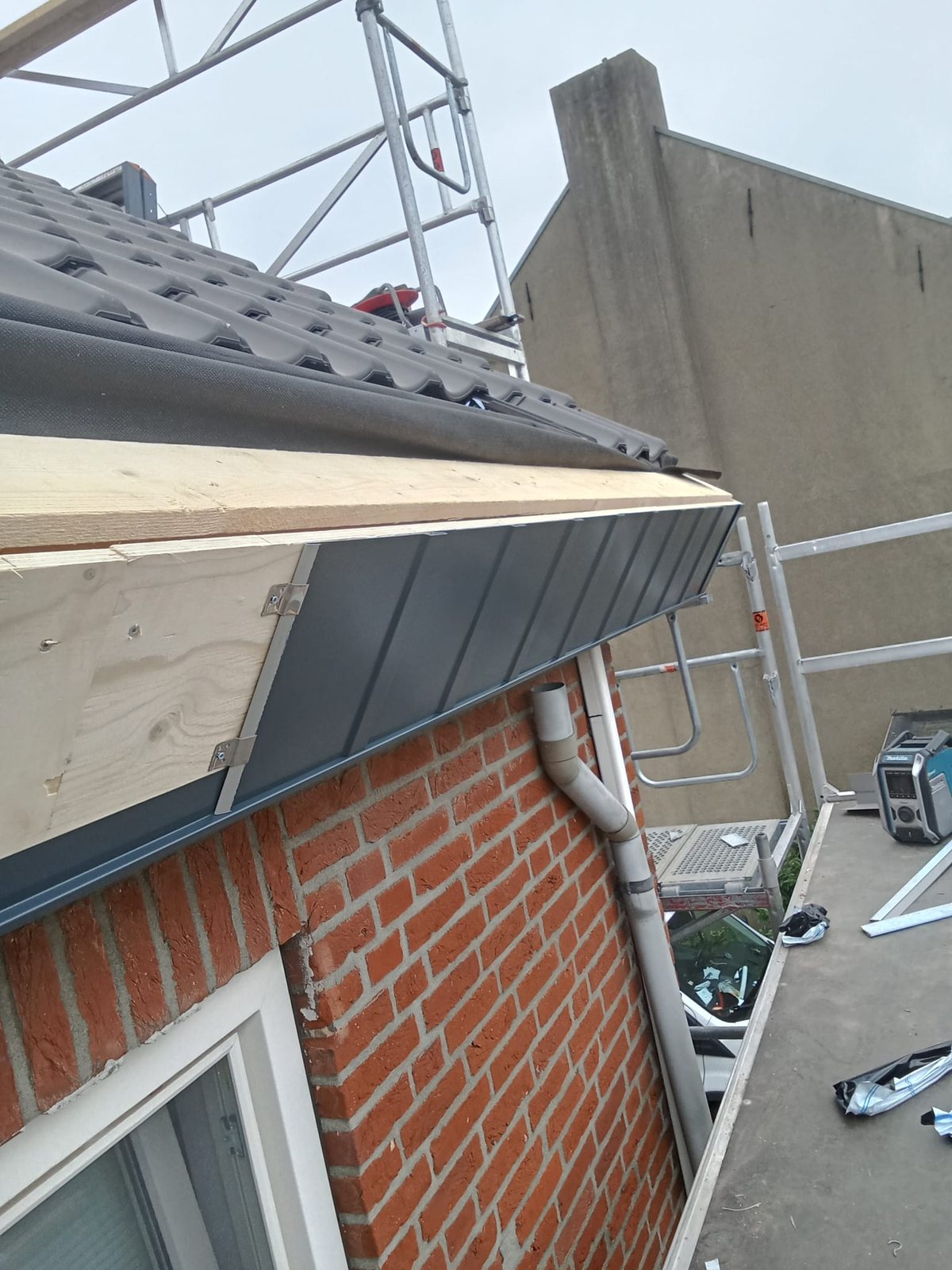 Nieuwe grijze boeiboordbekleding over vers hout aan de dakrand, met steiger en regenpijp