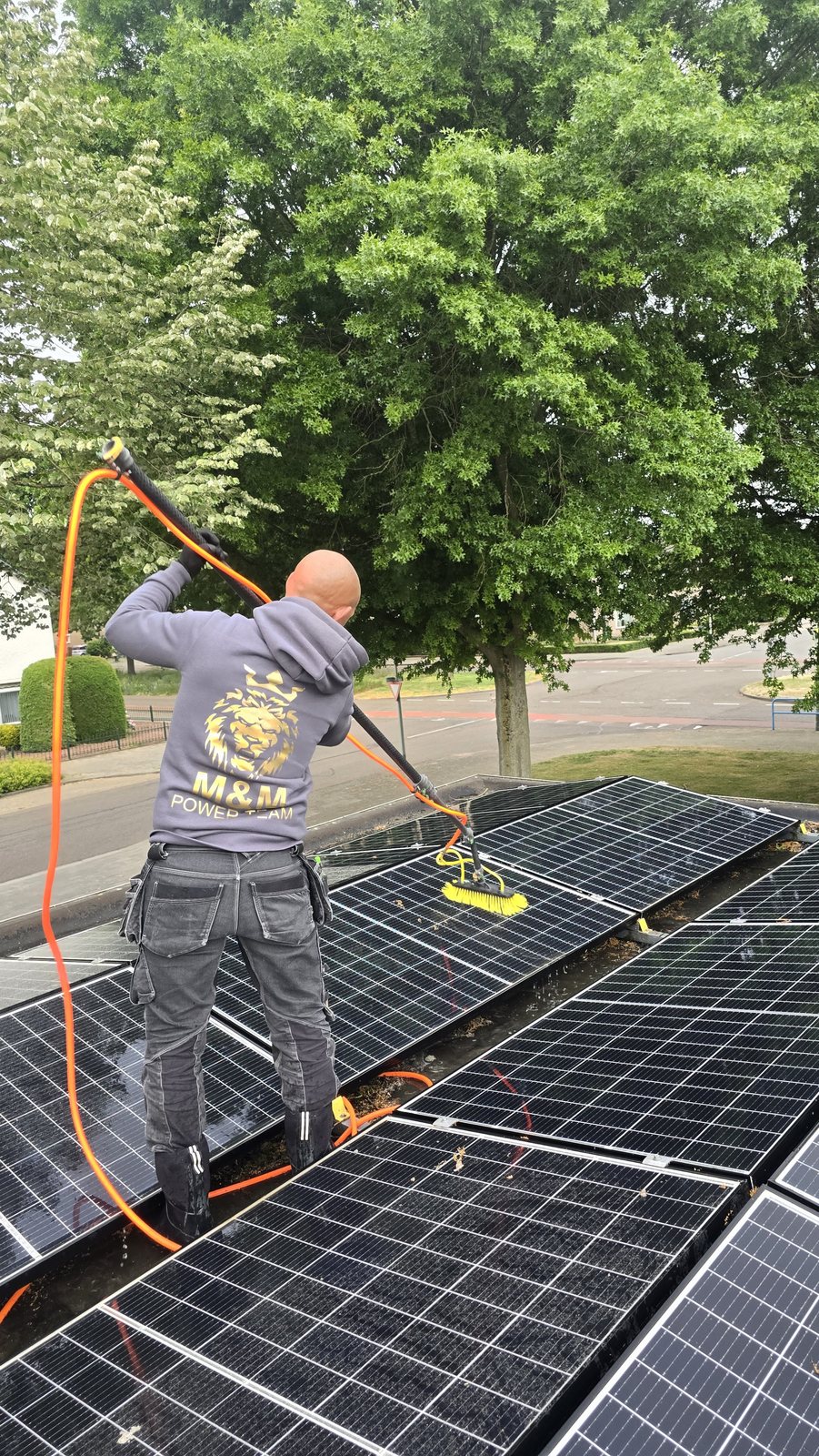 Teamlid in een hoodie met het M&M-logo reinigt zonnepanelen met een borstel op een waterslang